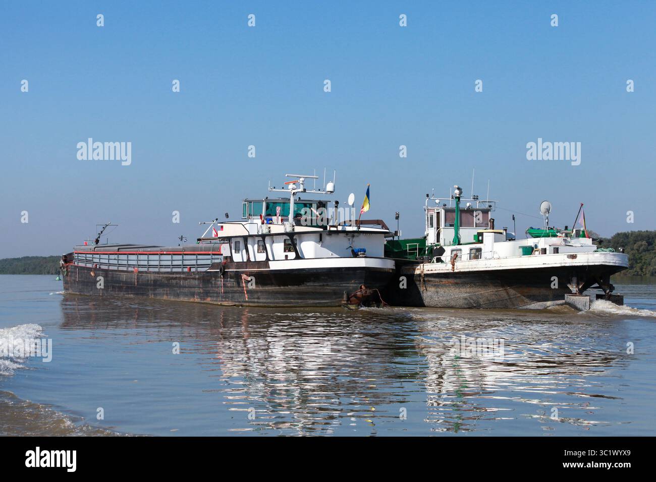 Zwei Frachtschiffe befahren die Donau unter klarem Himmel und spiegeln den modernen Verkehr auf Wasserstraßen vor einer ruhigen natürlichen Kulisse wider. Stockfoto