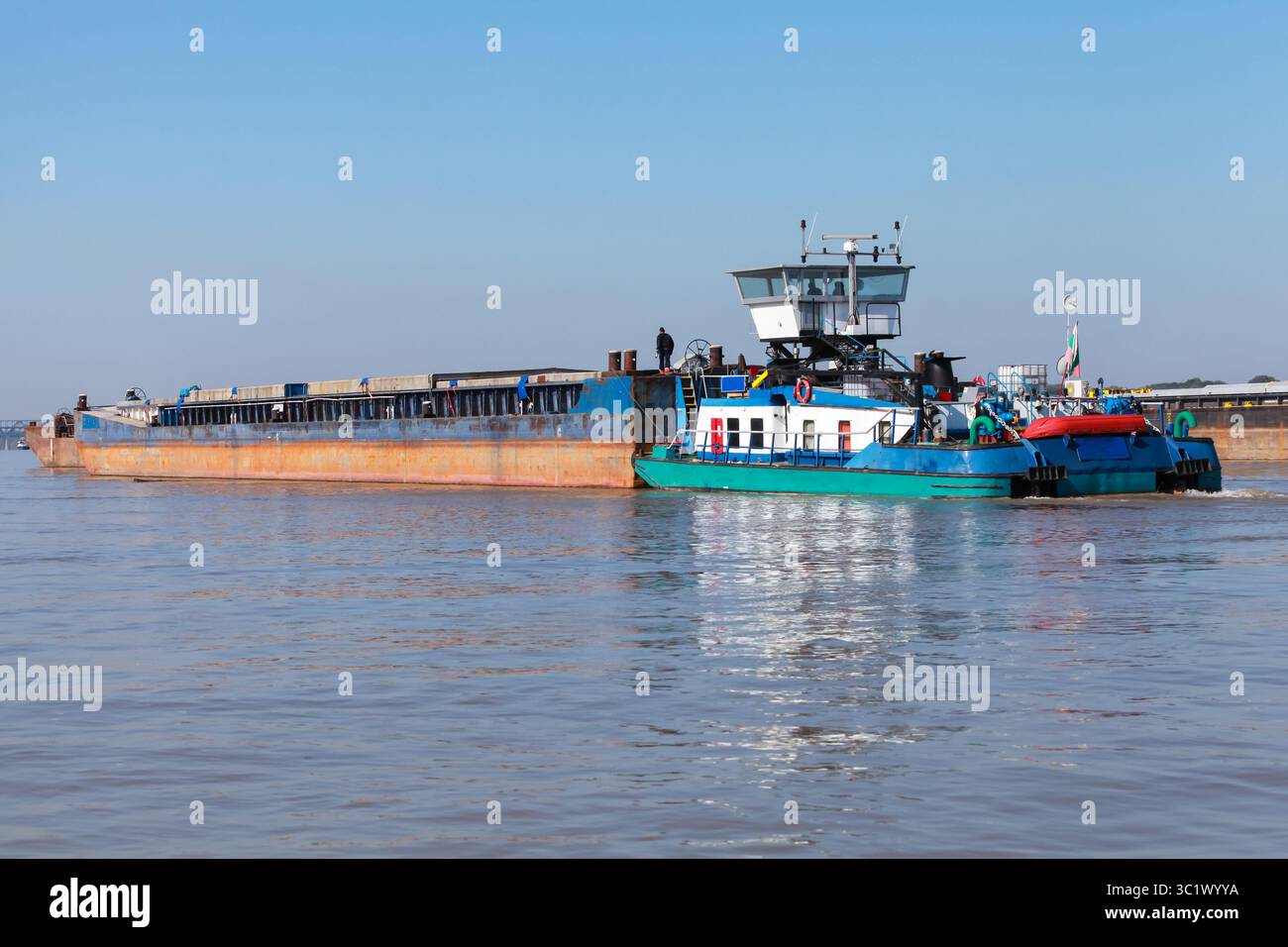 Rückansicht eines blau-weißen Schleppers, der auf einem Lastkahn auf der Donau fährt, mit einer Kulisse aus grünem Wald und einem klaren blauen Himmel, der sich auf dem Wasser spiegelt Stockfoto