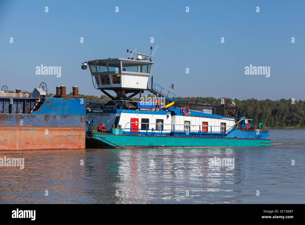 Ein blau-weißes Schlepper bewegt einen Lastkahn auf der Donau, mit einer Kulisse aus grünem Wald und einem klaren blauen Himmel, der auf dem Wasser reflektiert Stockfoto