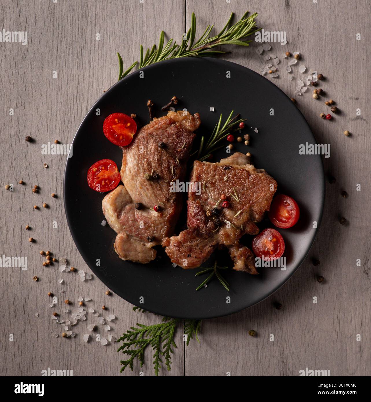 Eine schwarze Platte von oben nach unten mit gegrillten Lammkoteletts, Kirschtomaten, Rosmarinzweigen und Pfefferkörnern. Die Platte sitzt auf einem verwitterten grauen W Stockfoto