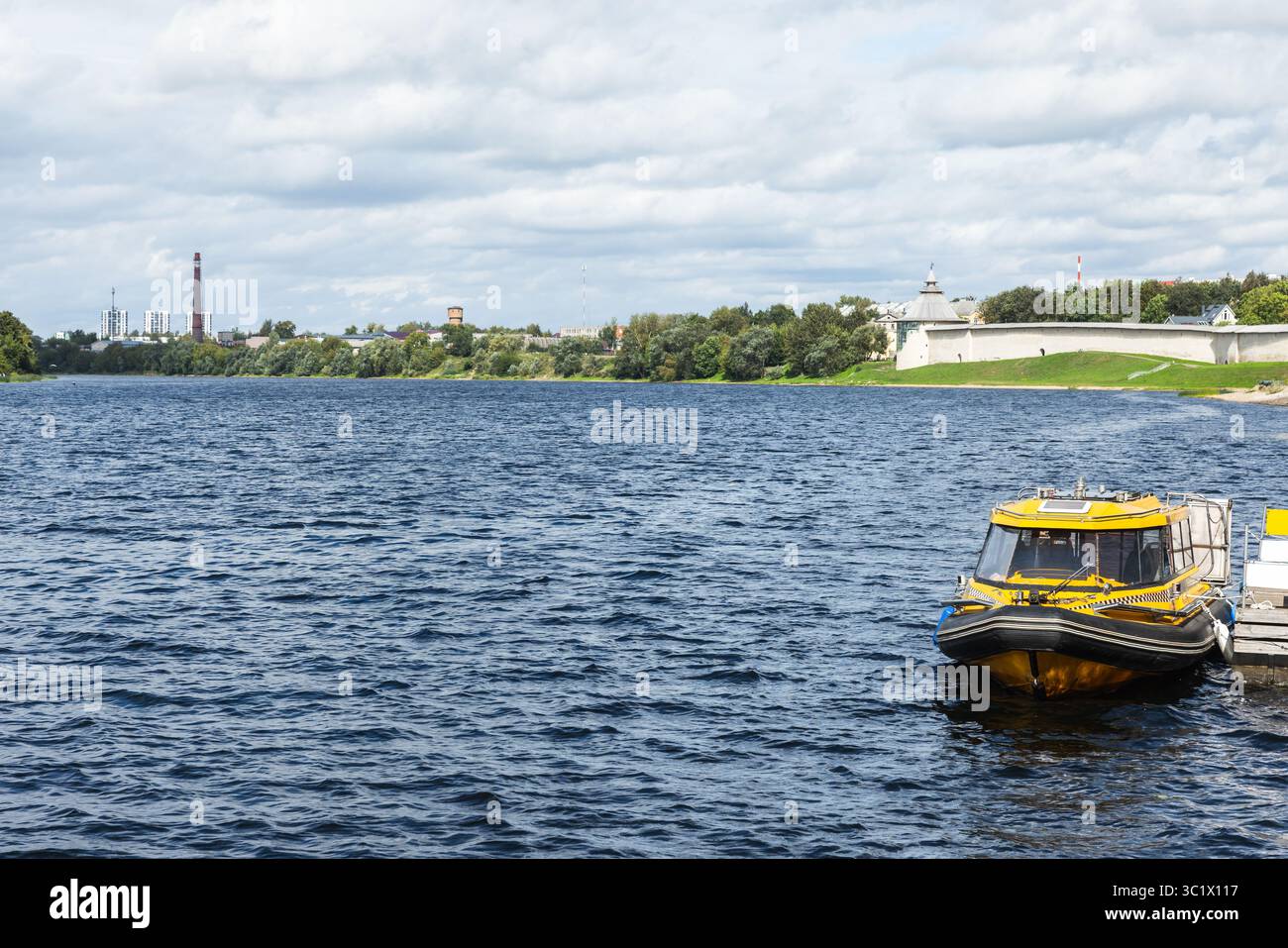 Ein lebhaftes gelbes Wassertaxi-Boot liegt friedlich an einem breiten blauen Fluss, mit einer historischen Steinfestung und üppigem Grün, das im Hinterland sichtbar ist Stockfoto