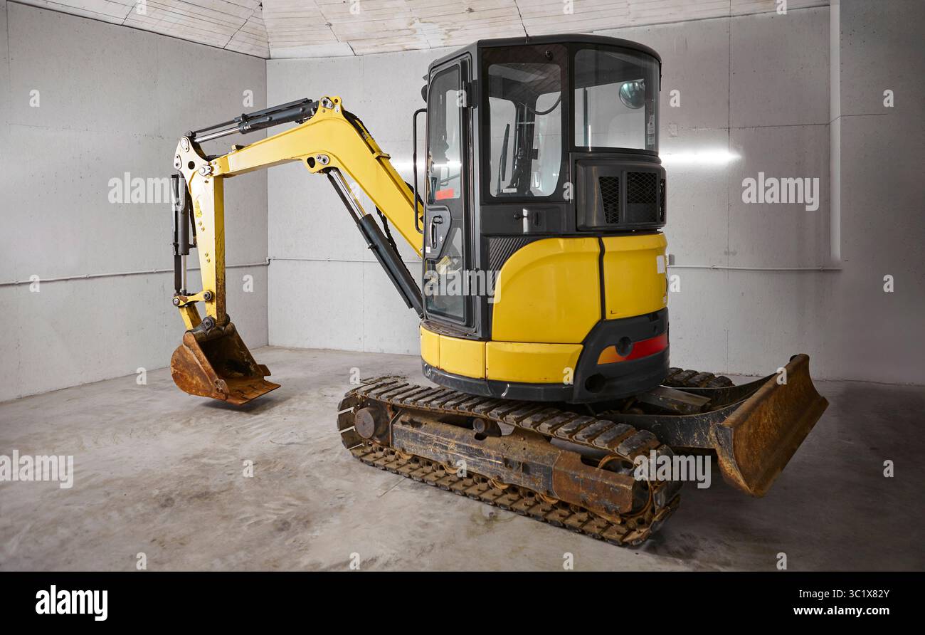 Ein gelber Minibagger befindet sich auf einem Betonboden in einem grauen, industriell aussehenden Raum. Der Bagger hat eine schwarze Kabine und einen hellgelben Arm und buc Stockfoto