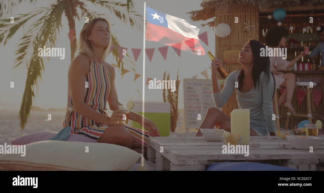 Lachende zwei Frauen, die Cocktails auf Palettentisch beim Sonnenuntergang am Strand halten, mit Tiki-Bar dahinter Stockfoto
