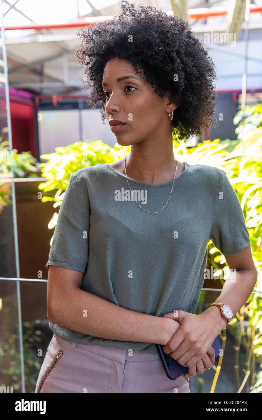 Afroamerikanische Frau, die in der Nähe eines Glasgeländes im Atrium steht, eine Armbanduhr mit Smartphone trägt Stockfoto