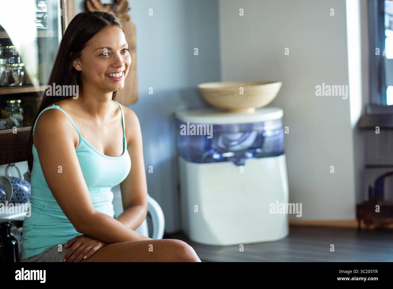 Frau sitzt in der Küche zwischen Regal mit Gläsern und Wasserspender mit Schüssel, Kopierraum Stockfoto