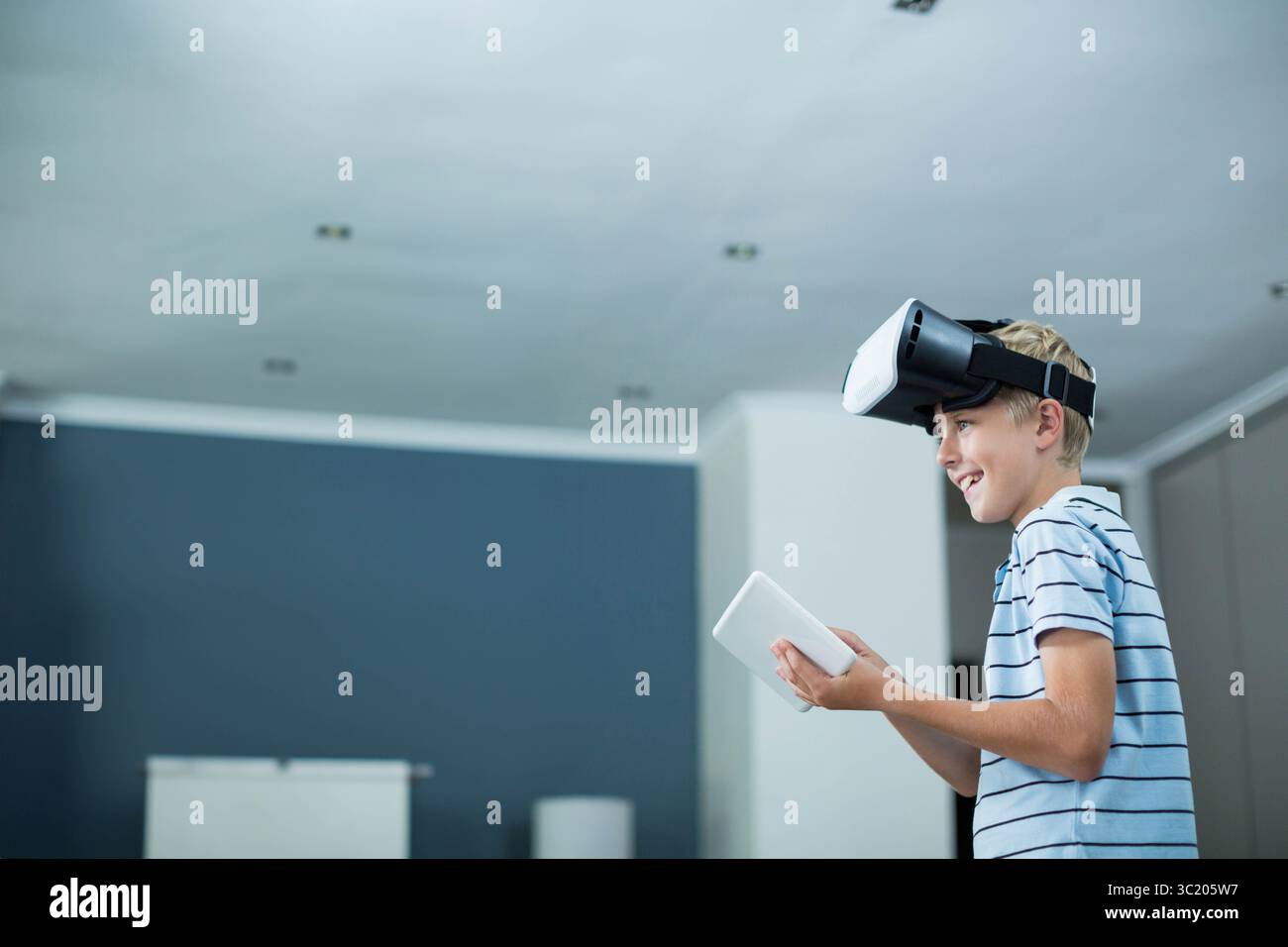 Männliches Kind, das in einem spärlich eingerichteten Raum steht und ein Tablet hält und das VR-Headset verwendet, Kopierraum Stockfoto