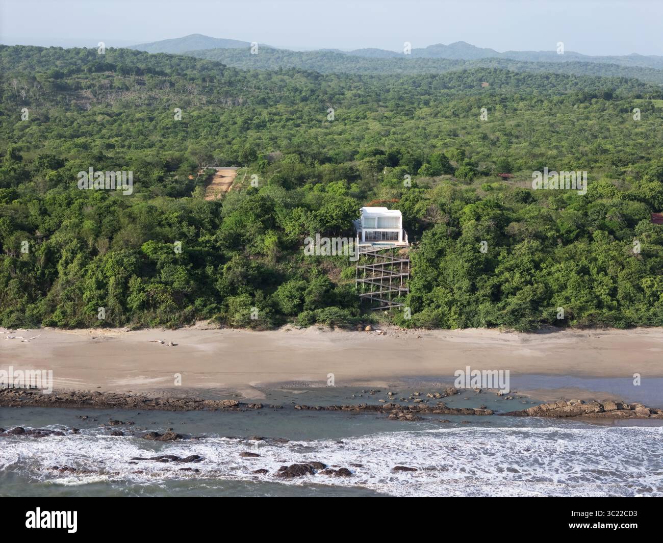 Das beeindruckende moderne Gebäude überblickt den Sandstrand, umgeben von dichtem Grün und Hügeln unter klarem Himmel. Stockfoto