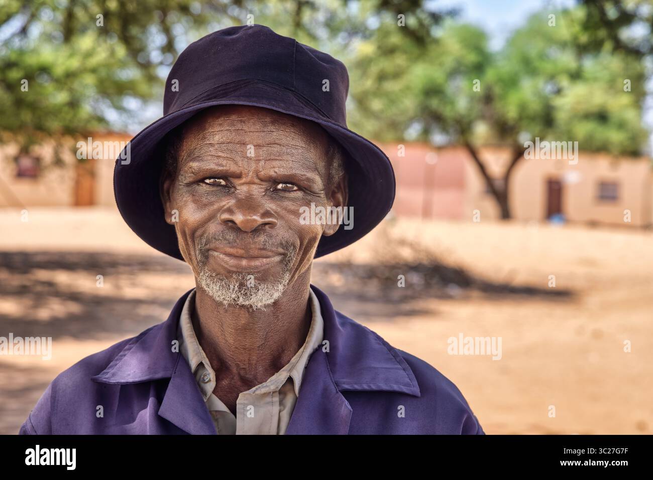 Porträt einzelner alter afrikanischer Mann im afrikanischen Dorf, Arbeitskleidung und ein Eimerhut, die Häuser in der Gemeinde sind sauber Stockfoto
