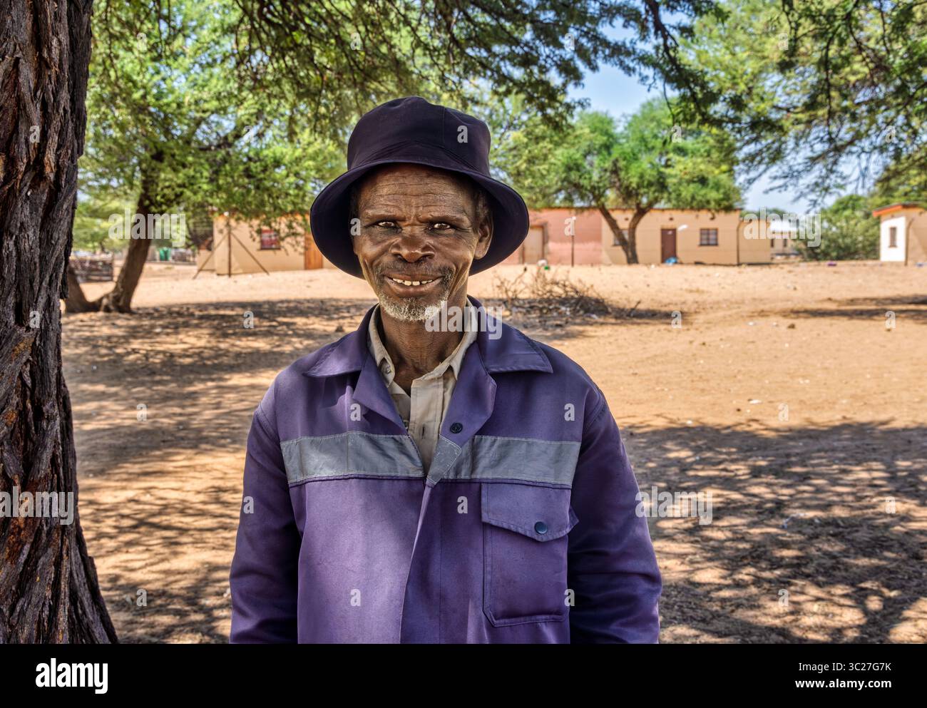 Porträt einzelner alter afrikanischer Mann im afrikanischen Dorf, Arbeitskleidung und ein Eimerhut, die Häuser in der Gemeinde sind sauber Stockfoto