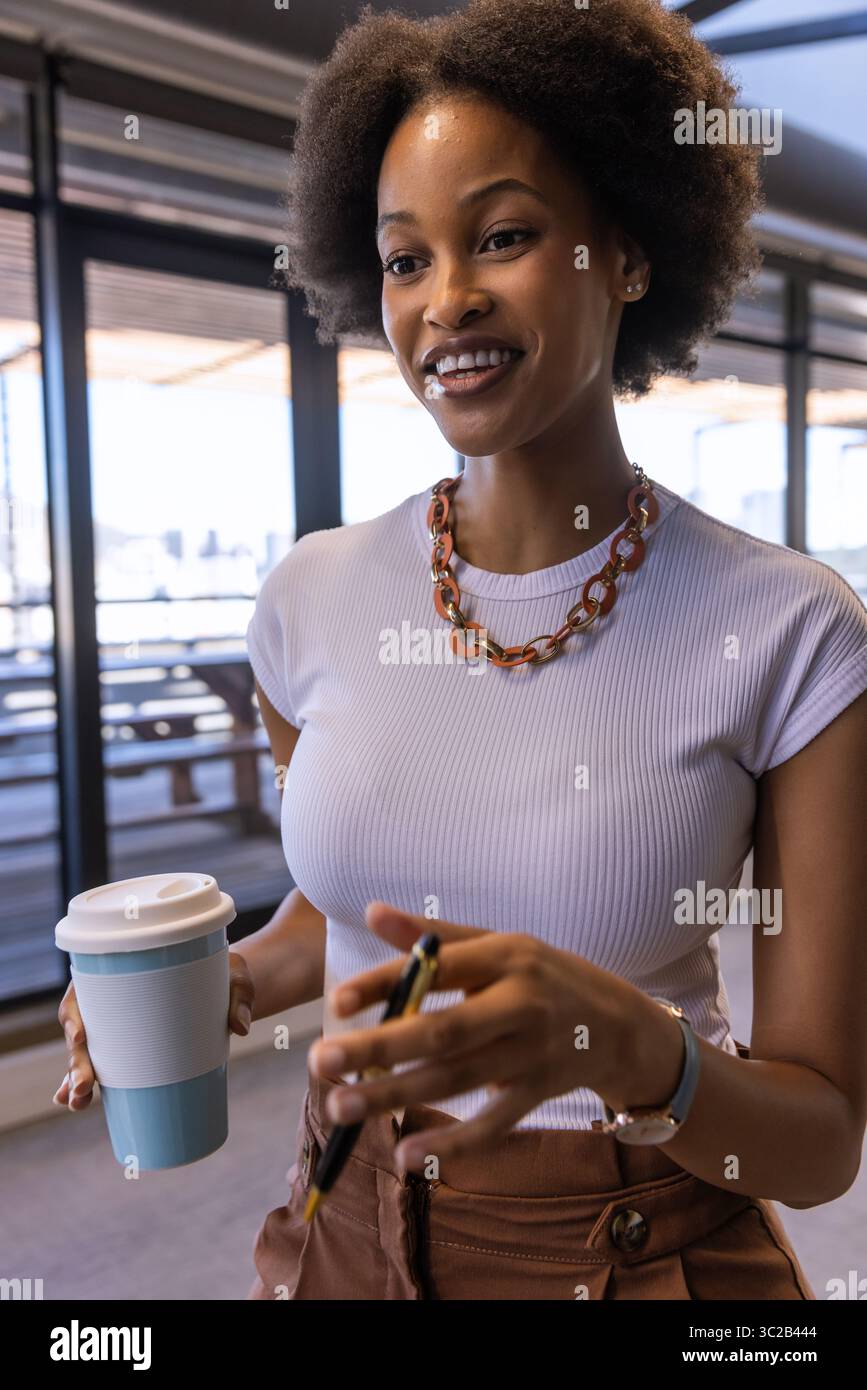Afroamerikanerin, die einen Reisebecher hält und ihr Smartphone in der Lounge in Geschäftskleidung überprüft Stockfoto