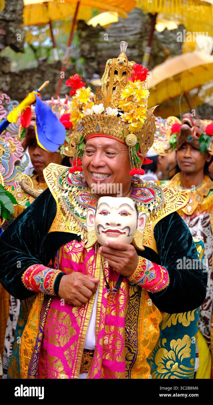 Balinesischer Mann in zeremonieller Tracht lächelt, während er eine traditionelle Maske hält Stockfoto