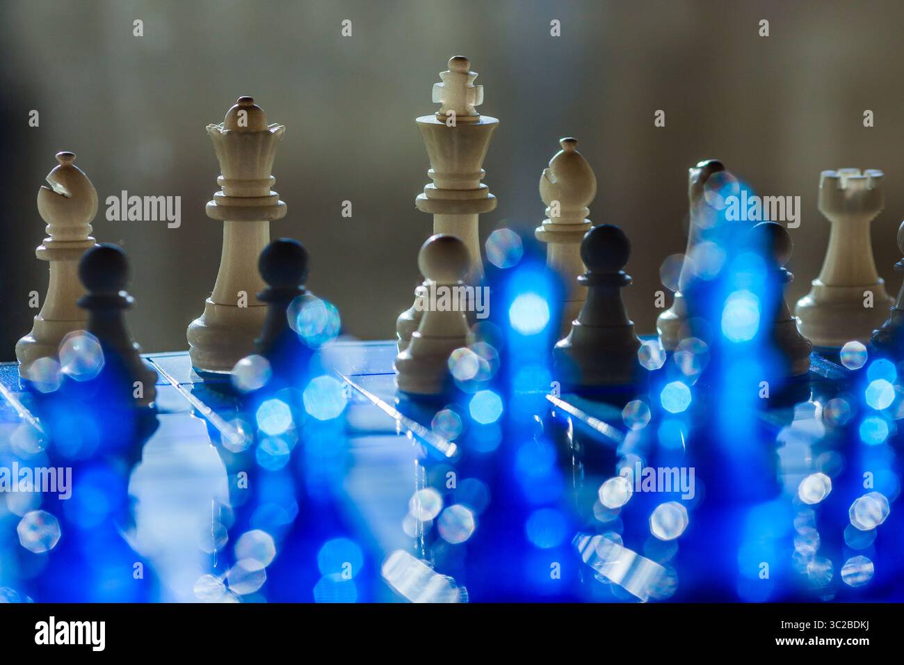 Eine dramatische Schachszene besteht aus Holzstücken auf reflektierender blauer Oberfläche und sanften Bokeh-Lichtern, die Tiefe und Intrige verleihen Stockfoto