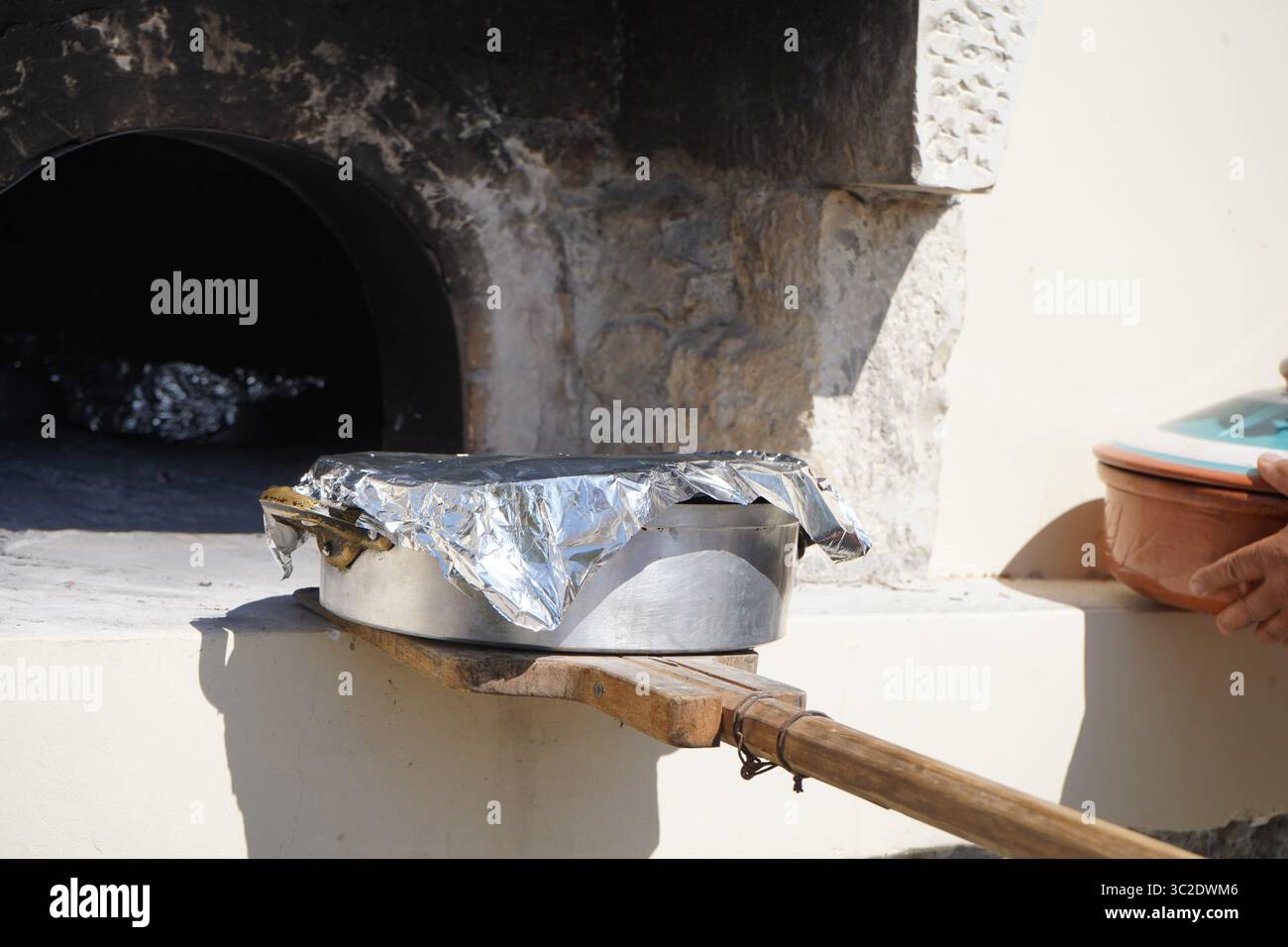 Der Mann verwendet einen langen Holzspachtel, um eine Folienschale in einen traditionellen Steinofen im Freien zu stellen. Ein keramisches Modell ist sichtbar. Stockfoto