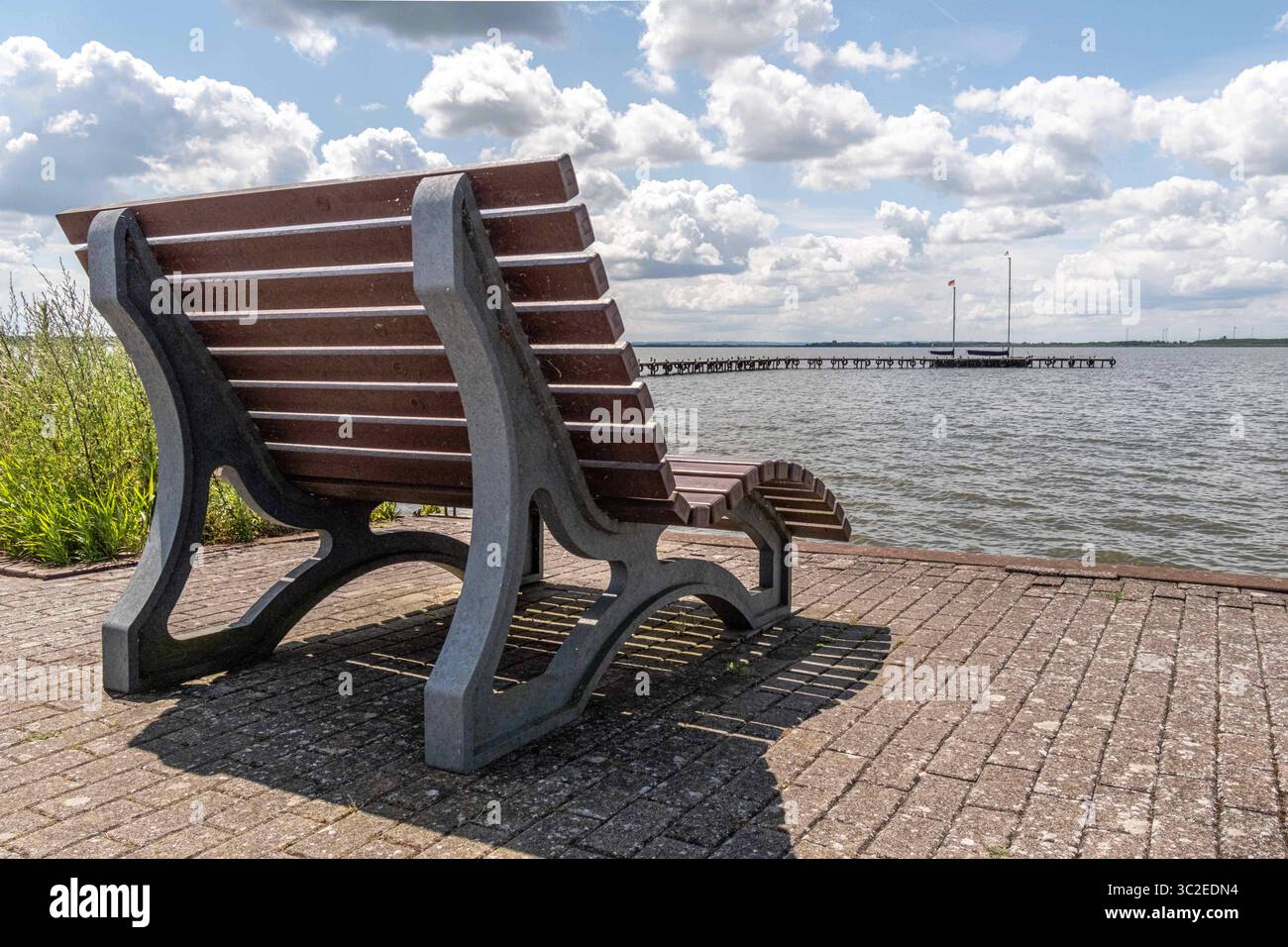 Dümmer, Deutschland 09. Juli 2025: Im Bild: Diese einladende Aufnahme zeigt eine moderne, robuste Sitzbank aus Holz und Stein, strategisch platziert auf einer gepflasterten Uferpromenade direkt am Dümmer See in Niedersachsen. Die Bank bietet einen malerischen Blick über die weite Wasserfläche des Sees, die unter einem strahlend blauen Himmel mit imposanten weißen Wolken liegt. Im Hintergrund erstreckt sich ein langer Bootssteg oder Pier weit in den See hinein, gesäumt von zahlreichen Segelbooten, was die Beliebtheit des Wassersports am Dümmer See unterstreicht. Diese Rastmöglichkeit lädt Besuc Stockfoto