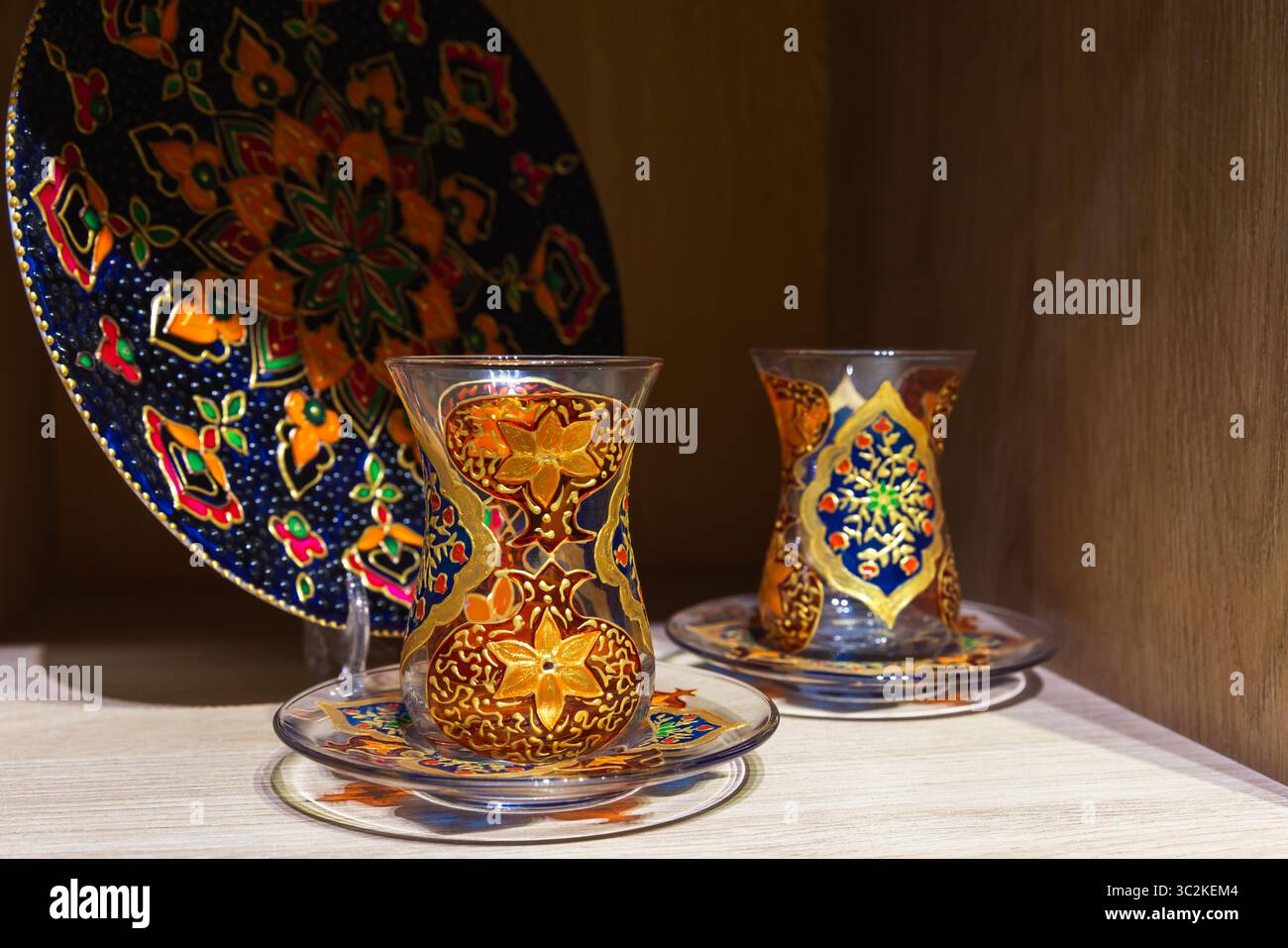 Aufwendig gestaltete handbemalte Teegläser Armudu mit einer farbenfrohen Keramikplatte, die auf einem Holzregal ausgestellt ist und die traditionelle Handwerkskunst unterstreicht Stockfoto