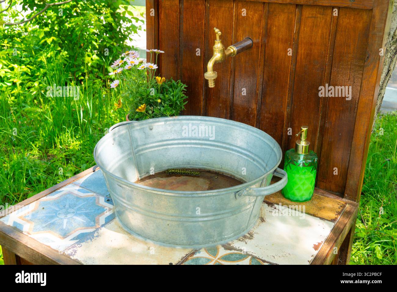 Ein rustikaler Außenwaschtisch mit einem Vintage-Messinghahn, einem Zinkeimer und einem grünen Seifenspender, der auf einer gefliesten Oberfläche in einem sonnigen Garten sitzt Stockfoto