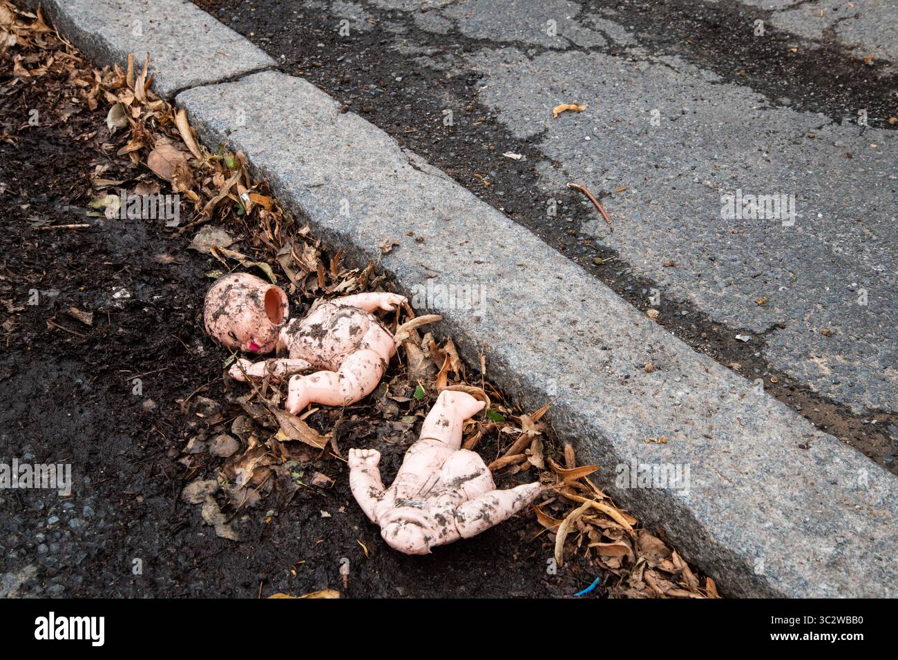 Die kaputte und dreckige Babypuppe liegt am Straßenrand zwischen toten Blättern und Schlamm. Eine eindringliche, urbane Verfallsszene, die Vernachlässigung, verlorene Unschuld symbolisiert Stockfoto