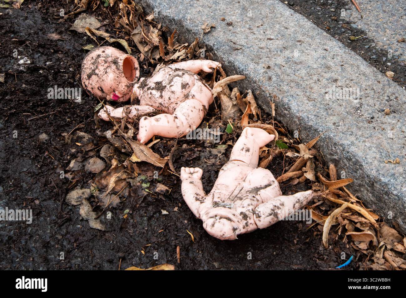 Die kaputte und dreckige Babypuppe liegt am Straßenrand zwischen toten Blättern und Schlamm. Eine eindringliche, urbane Verfallsszene, die Vernachlässigung, verlorene Unschuld symbolisiert Stockfoto