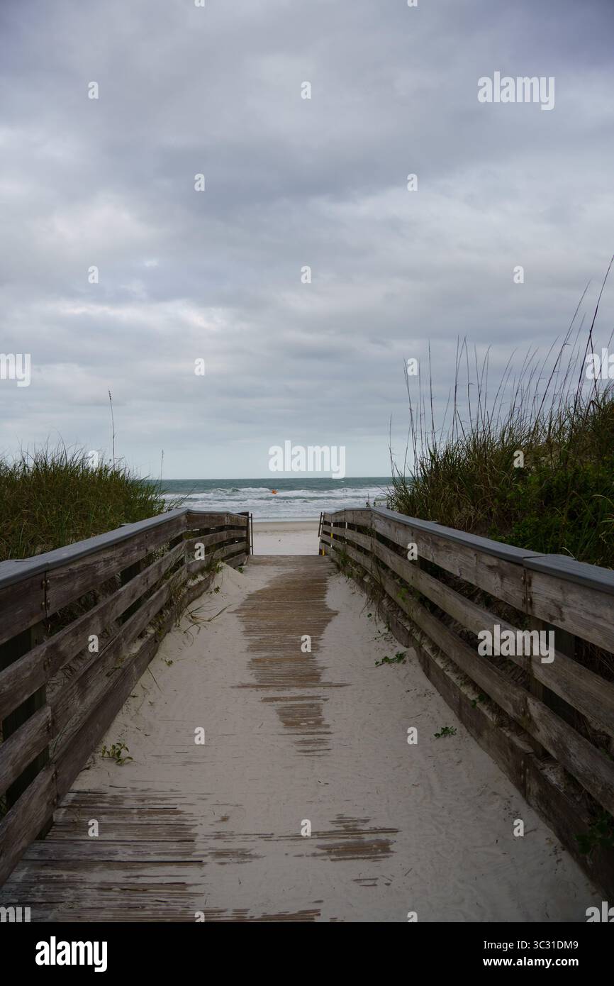 Hölzerner Fußweg, der an einem bewölkten Tag durch Dünengras zur Küste führt. Stockfoto