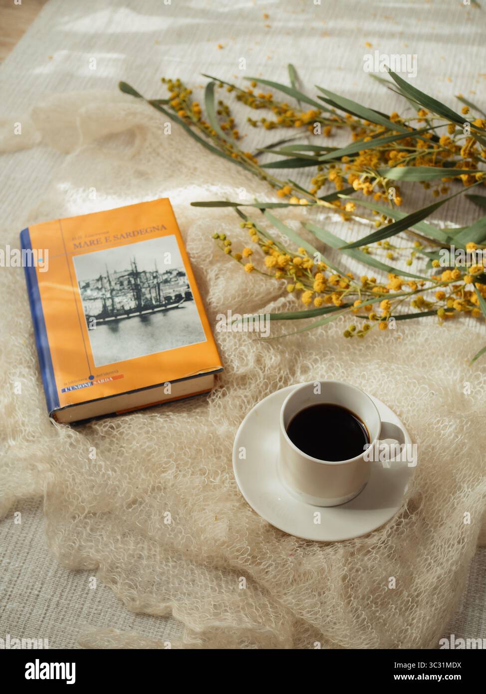 Italienische Ausgabe des Romans „Meer und Sardinien“ („Mare e Sardegna“) von D.H. Lawrence mit Mimosa und Kaffee auf weißem Kaschmirschal – vertikale Nahaufnahme, ol Stockfoto