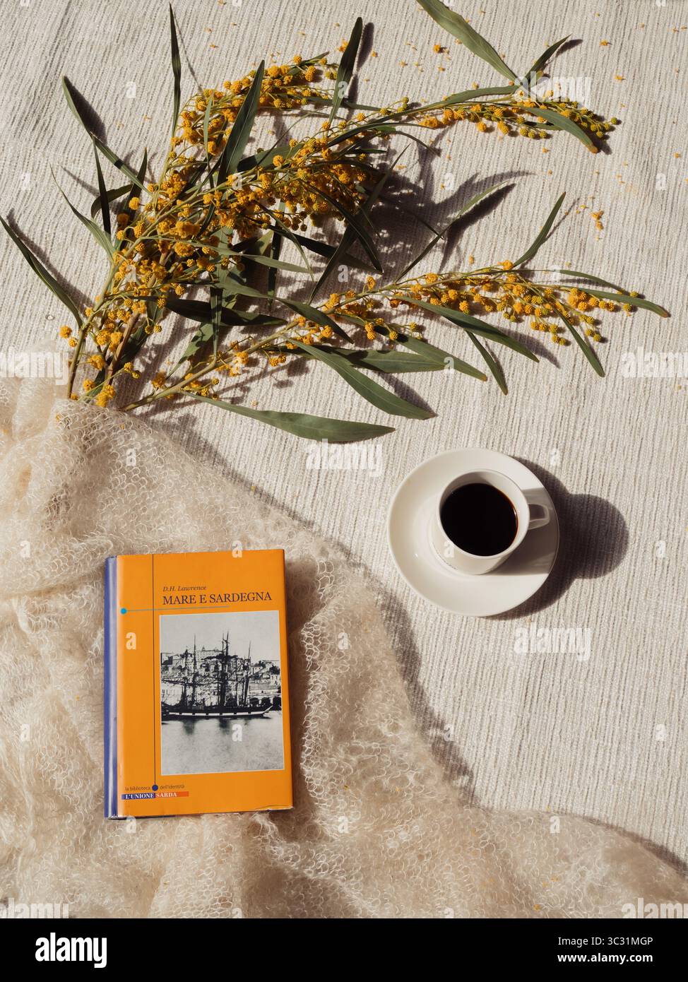Italienische Ausgabe des Romans „Meer und Sardinien“ („Mare e Sardegna“) von D.H. Lawrence mit Mimosa und Kaffee auf weißem Kaschmirschal – vertikale Nahaufnahme, ol Stockfoto