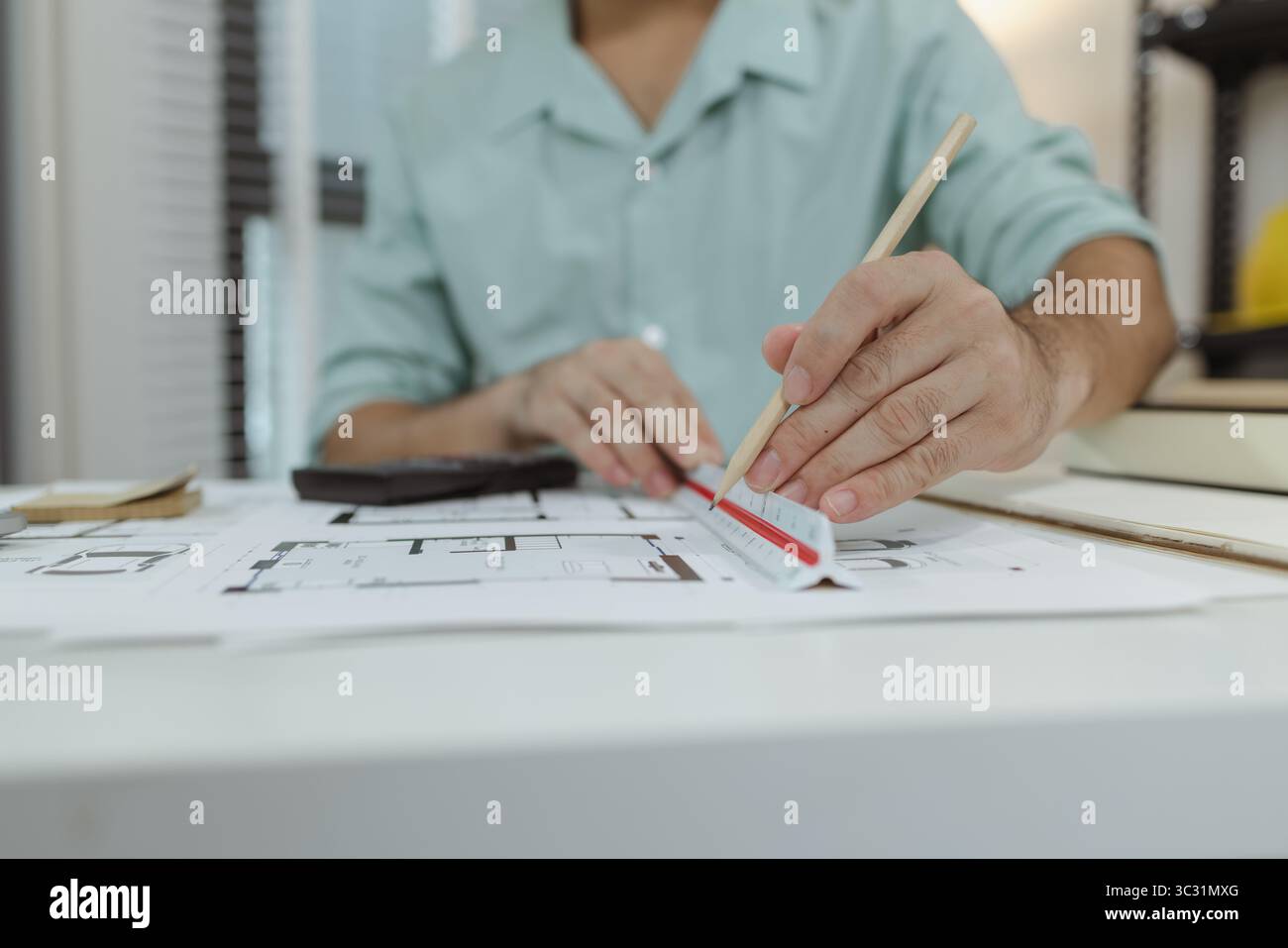 Ein Architekt, der an einem weißen Schreibtisch mit Blaupausen, einem Taschenrechner und einem dreieckigen Maßstabslineal arbeitet und sich auf das Zeichnen und Messen von Det konzentriert Stockfoto