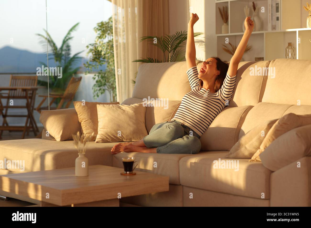 Aufgeregte und glückliche Frau, die die Arme hebt und zu Hause bei Sonnenuntergang auf einer Couch sitzt Stockfoto