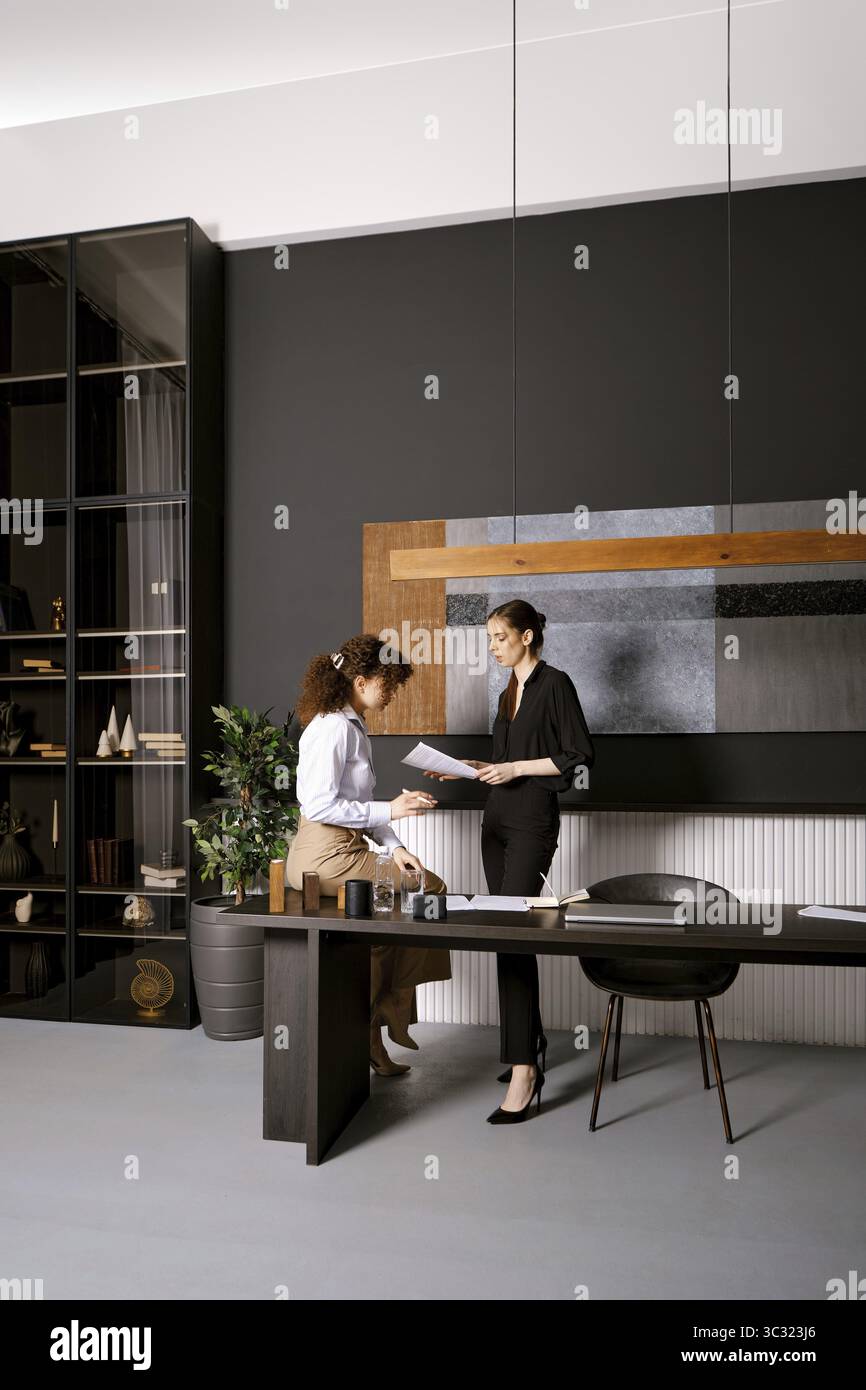 Zwei Geschäftsfrauen suchen nach einer Lösung für ein Problem, das in einem Büro Papiere liest. Eine Frau steht an einem schlanken Schreibtisch und überprüft Dokumente, während die o Stockfoto