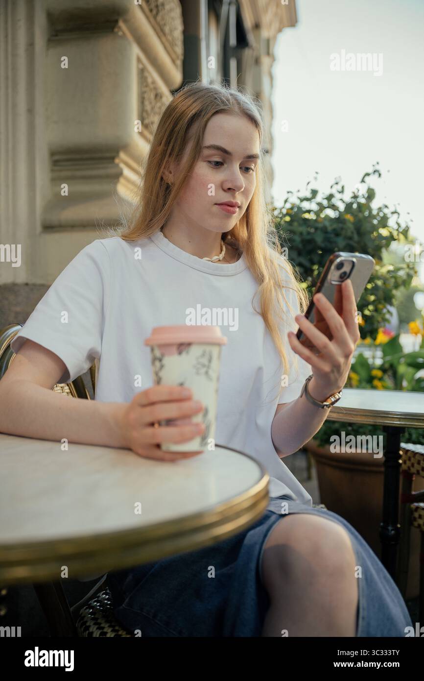 Junge Frau trinkt Kaffee im Café im Freien und benutzt Smartphone Stockfoto