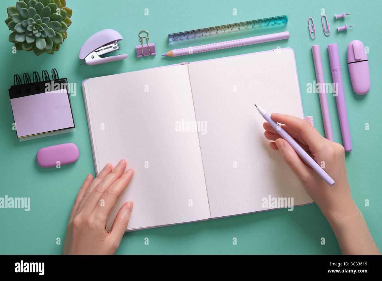 Leere Notizblock-Seite für Ihren Text in einem pastellfarbenen Briefpapierrahmen. Die Hände der Frau halten einen Stift. Hintergrund in mintgrüner Farbe. Stockfoto