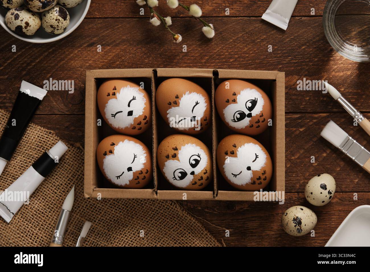 Handbemalte Eulen-Ostereier in einer Schachtel. Umgeben von Malutensilien auf hölzernem Hintergrund. Kreative Weihnachtsdekoration Stockfoto