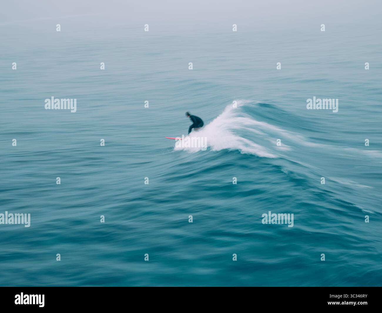 Eine einsame, verschwommene Surferfigur, die sich zwischen den Wellen in einer blauen Meereslandschaft mit Bewegung verloren hat Stockfoto
