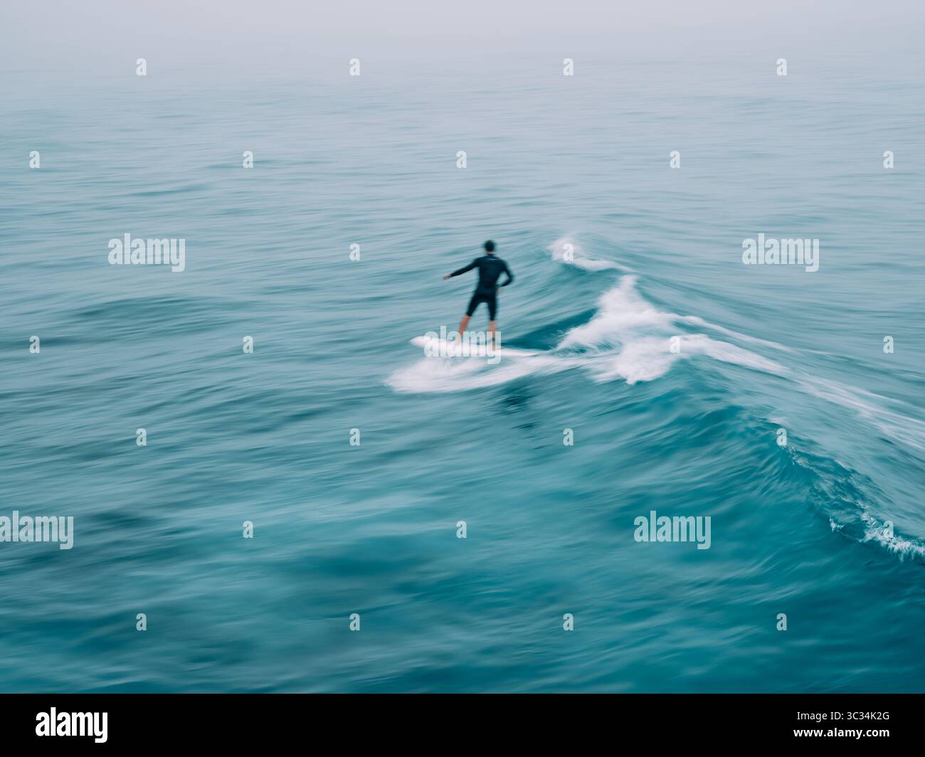 Eine einsame, verschwommene Surferfigur, die sich zwischen den Wellen in einer blauen Meereslandschaft mit Bewegung verloren hat Stockfoto