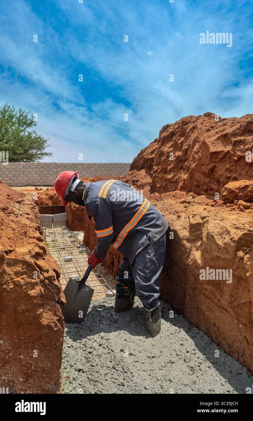 Arbeiter Afroamerikaner mit Schaufel ausgestattet, Helm und Uniform, die ein Betonfundament in einem Graben, umgeben von Erdhaufen, darstellen Stockfoto
