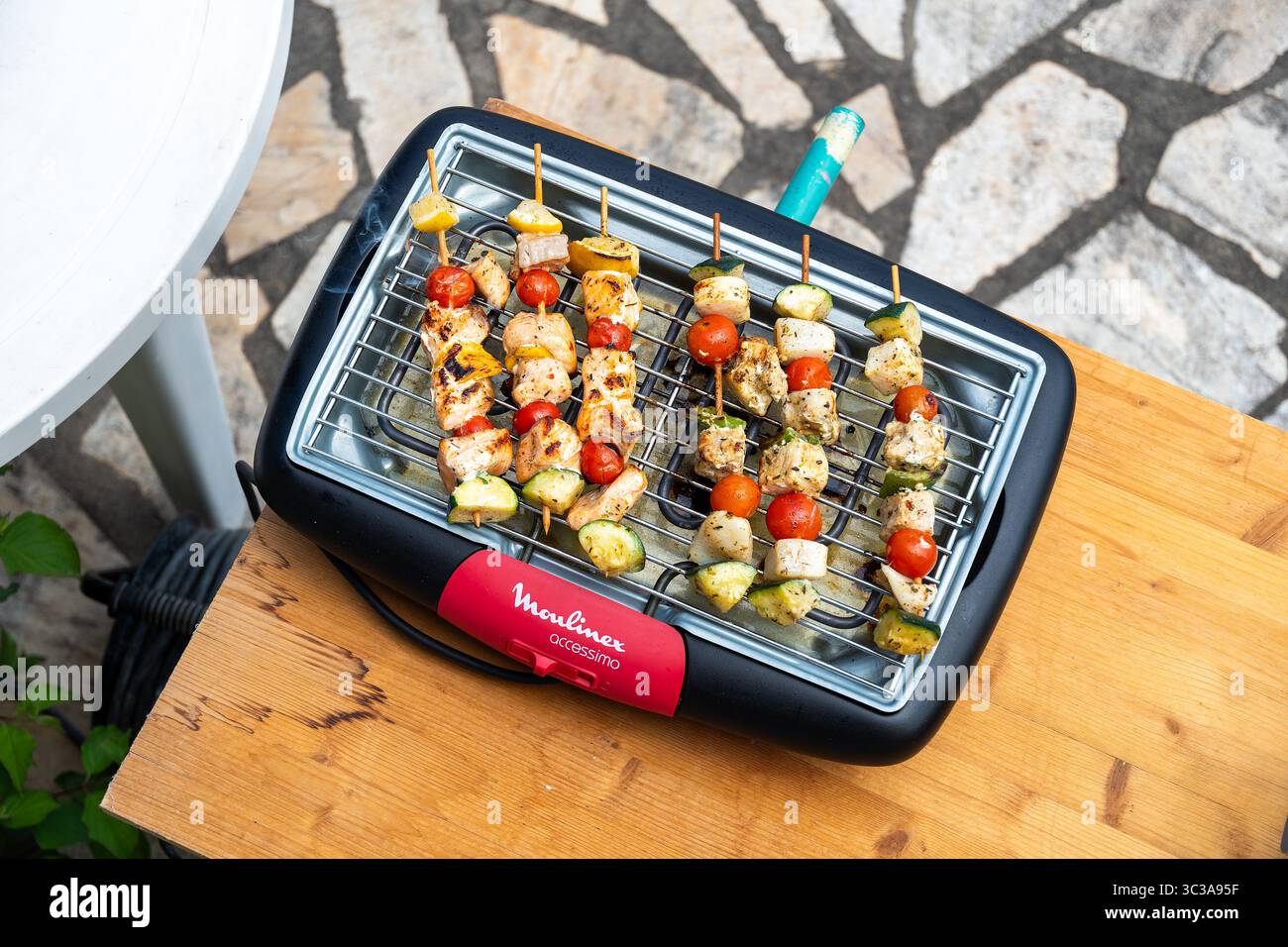 Eine Reihe hausgemachter Hühnchen- und Gemüsespieße, die auf einem elektrischen Moulinex-Grill grillen, auf einem Holztisch im Freien. Ein gesundes und farbenfrohes Di Stockfoto