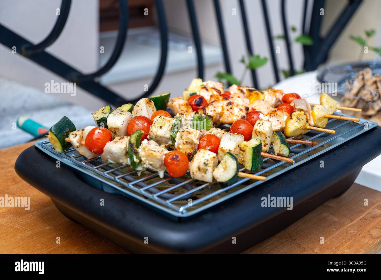 Eine Reihe hausgemachter Hühnchen- und Gemüsespieße, die auf einem elektrischen Moulinex-Grill grillen, auf einem Holztisch im Freien. Ein gesundes und farbenfrohes Di Stockfoto