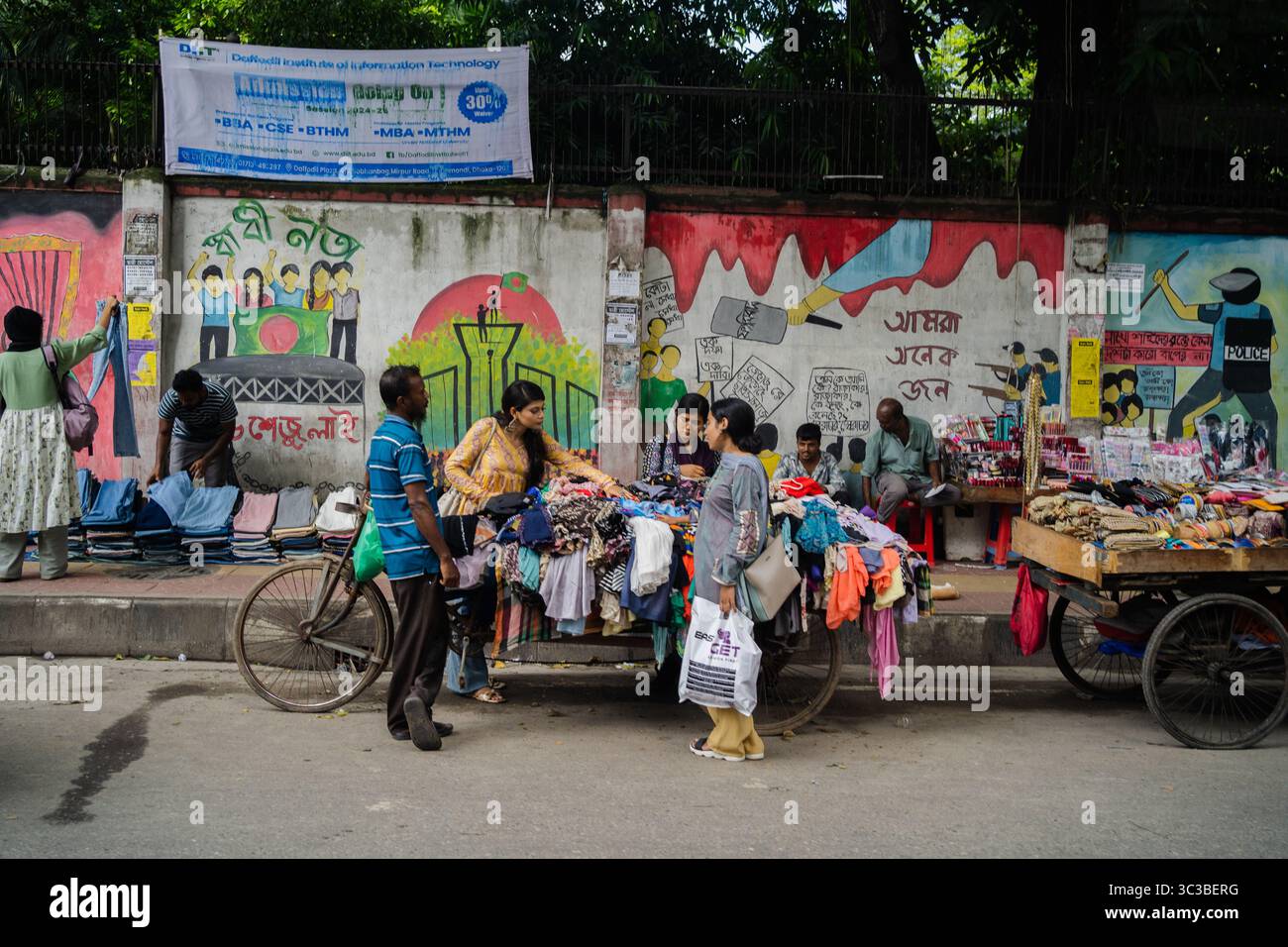 Eine lebhafte Straßenmarktszene in Dhaka, Bangladesch, wo ein Händler Kleidung direkt vom Fahrrad verkauft, neben anderen Verkaufsständen. Stockfoto