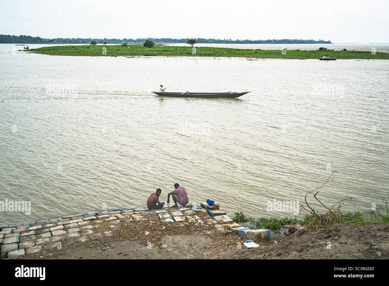 Ruhiges Leben am Flussufer im ländlichen Bangladesch – zwei Männer ruhen am Jamuna River, während ein traditionelles Boot vorbeifährt Stockfoto