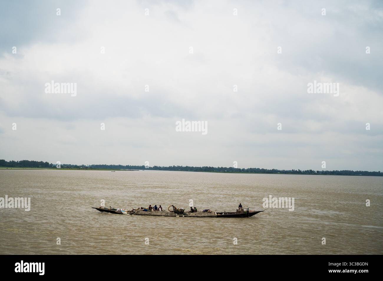 Ruhiges Leben am Flussufer im ländlichen Bangladesch – zwei Männer ruhen am Jamuna River, während ein traditionelles Boot vorbeifährt Stockfoto