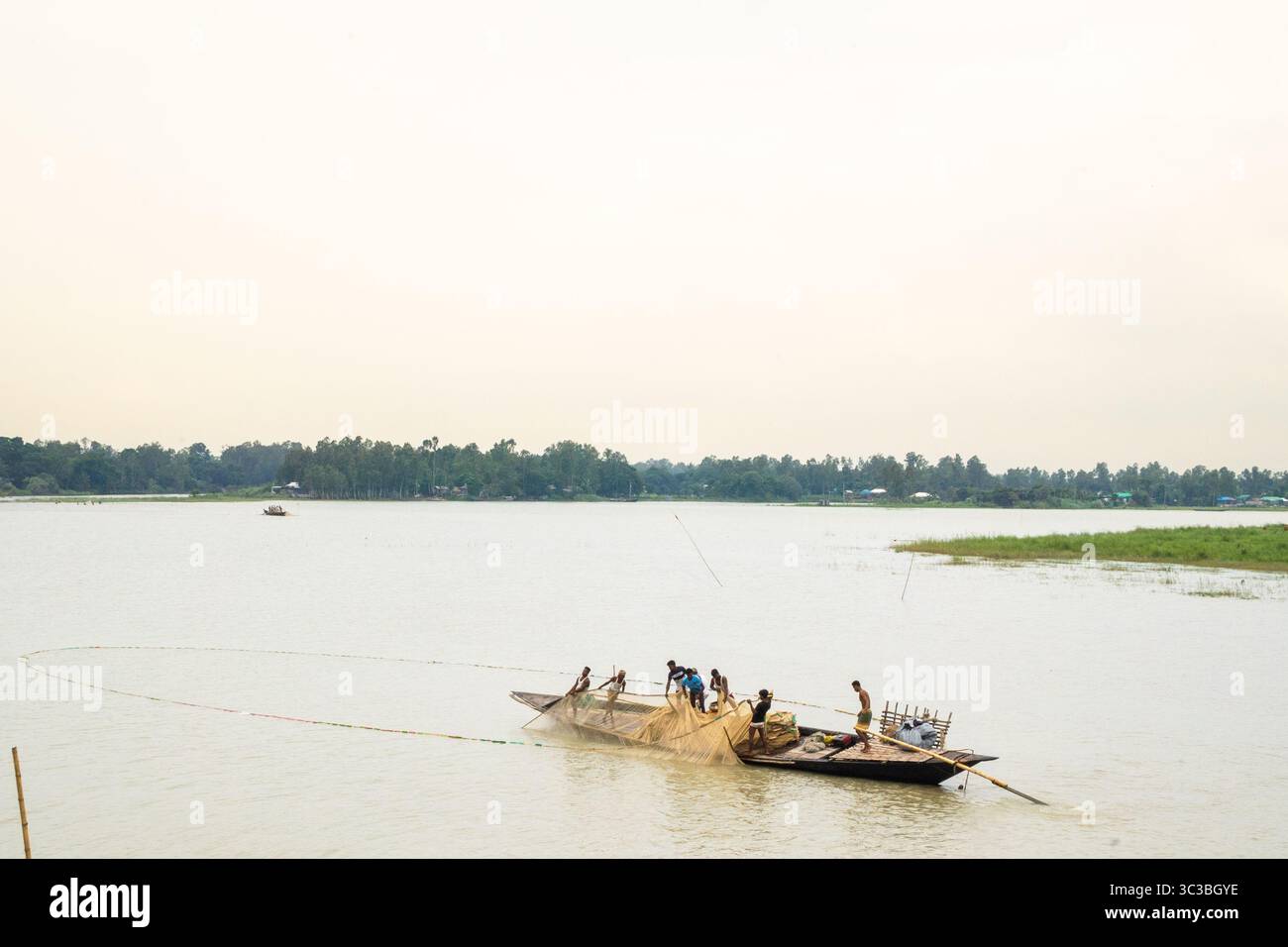 Ruhiges Leben am Flussufer im ländlichen Bangladesch – zwei Männer ruhen am Jamuna River, während ein traditionelles Boot vorbeifährt Stockfoto