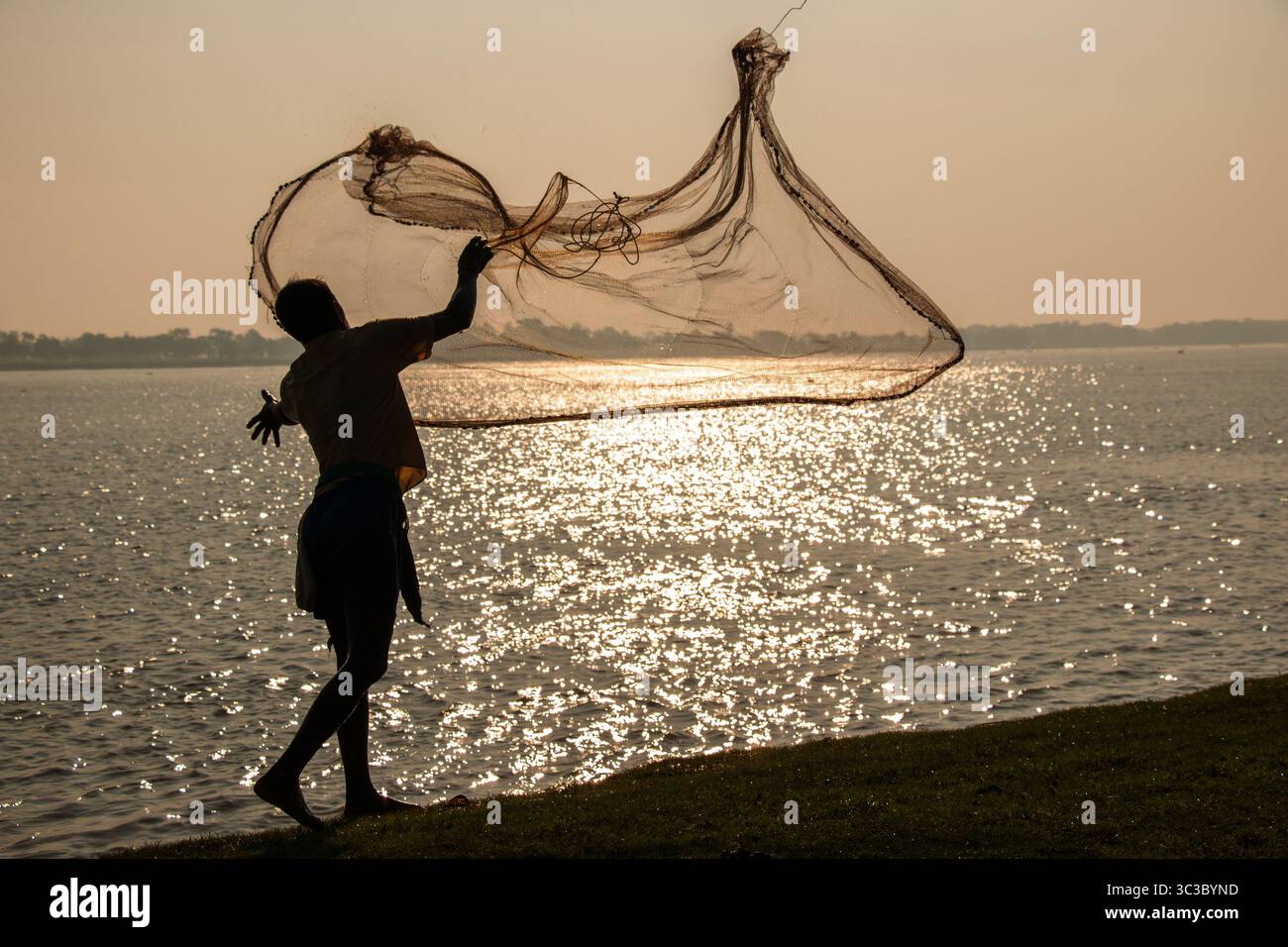 Ein Fischer wirft morgens sein Netz auf einen ruhigen Fluss im ländlichen Bangladesch und fängt den zeitlosen Rhythmus des Lebens und der Tradition ein. Stockfoto