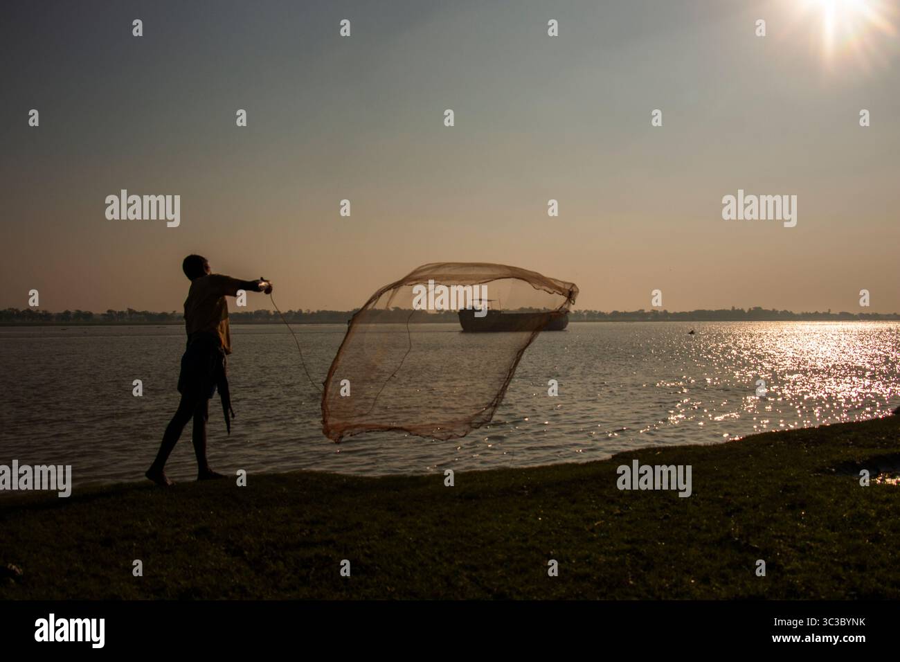 Ein Fischer wirft morgens sein Netz auf einen ruhigen Fluss im ländlichen Bangladesch und fängt den zeitlosen Rhythmus des Lebens und der Tradition ein. Stockfoto
