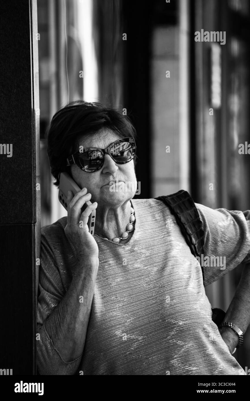 Reife Frau, die Sonnenbrille trägt und über Handy in der städtischen Umgebung spricht. Stockfoto