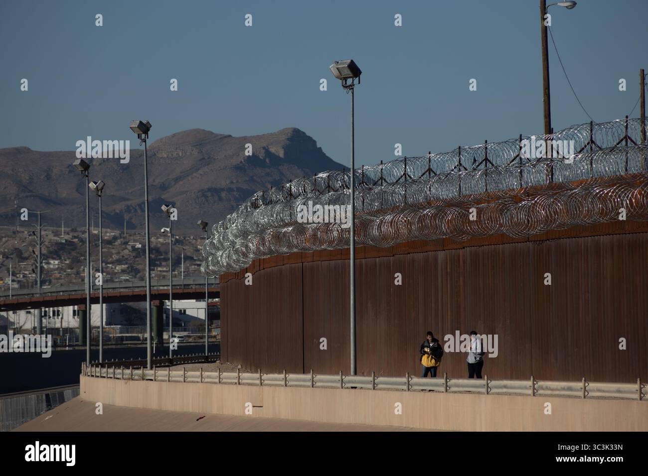 Zwei Menschen gehen am befestigten Grenzzaun in Ciudad Juarez vorbei und zeigen die Herausforderungen der Migration auf. Stockfoto