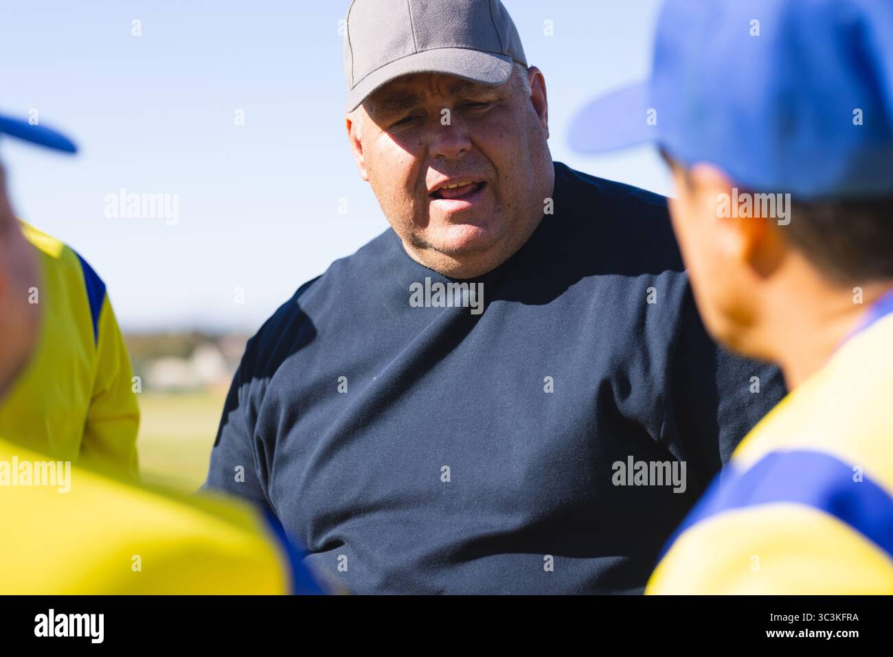 Männlicher Trainer, der Spieler in gelben Trikots auf dem Grasfeld mit Baseballmütze und marineblauem Hemd anweist Stockfoto
