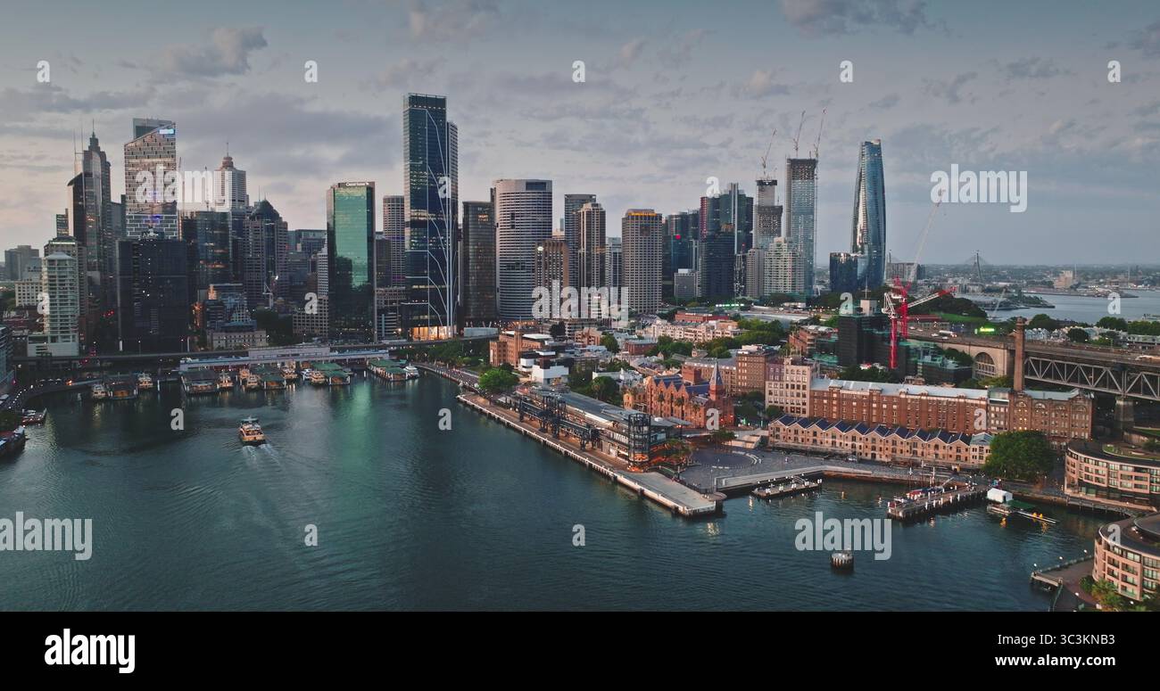 Australien, Sydney: CBD Skyline reflektiert auf ruhigem Wasser bei bewölktem Sonnenuntergang, moderne Gebäude mit Stadtblick Wolkenkratzer überraschen über Fährfahrten und Segeln im Hafen. Panorama der Drohne aus der Vogelperspektive Stockfoto