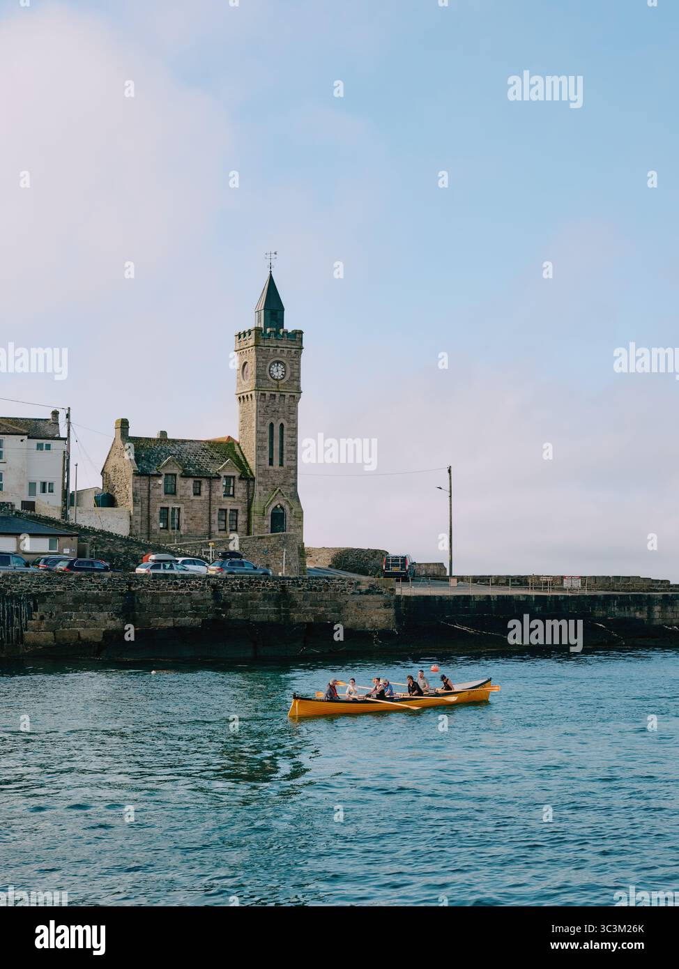 Die Hafenmauer und die Kirche mit vorbeiziehender Ruderclub-Gig-Crew in Porthleven, einem Fischerhafen in Cornwall, England. UK Stockfoto