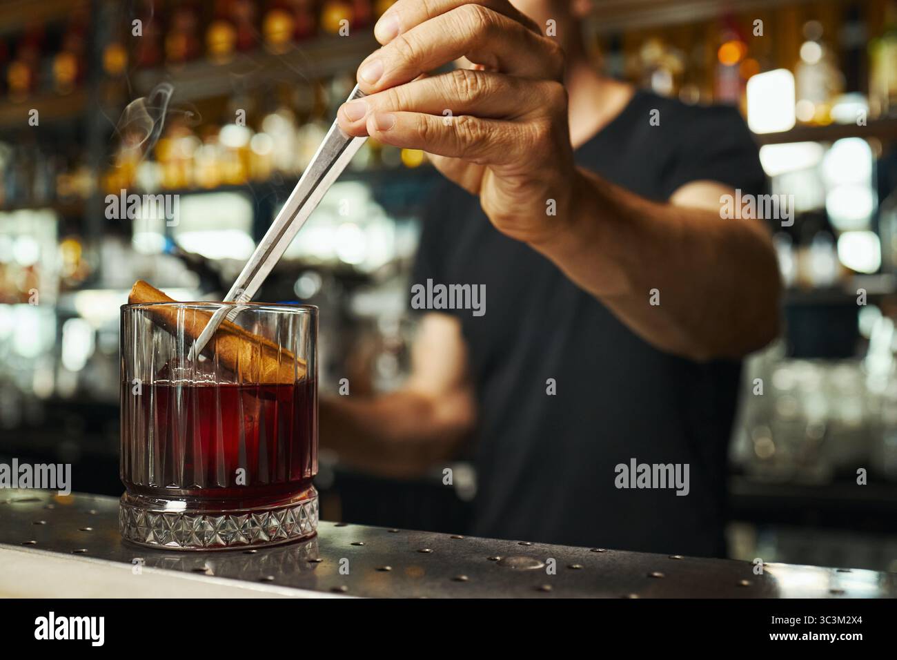 Kaukasischer junger Erwachsener, der Cocktail mit Zimtstab im Glas mit Metall-Barlöffel rührt, steht hinter der Bar im Restaurant mit Flaschenregalen im Hintergrund Stockfoto