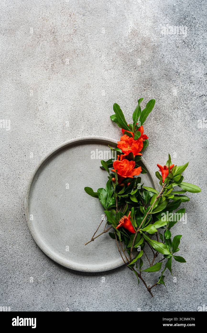 Ein Granatapfelzweig mit leuchtenden roten Blüten ist elegant auf einer einfachen runden Platte angeordnet, die vor einem strukturierten Hintergrund eine markante nat bildet Stockfoto