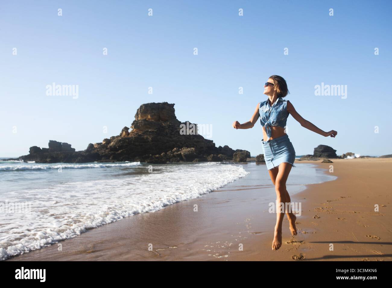 Die modische, schlanke junge Frau mit femininem Denim-Outfit läuft am Strand vor Felsformationen und dem Meer in Portugal. Vergnügen, Reisen Stockfoto